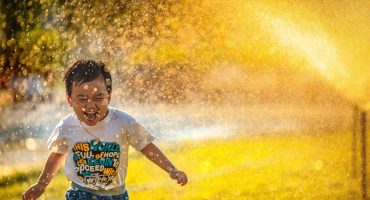 A young child runs through sprinklers in warm yellow light.
